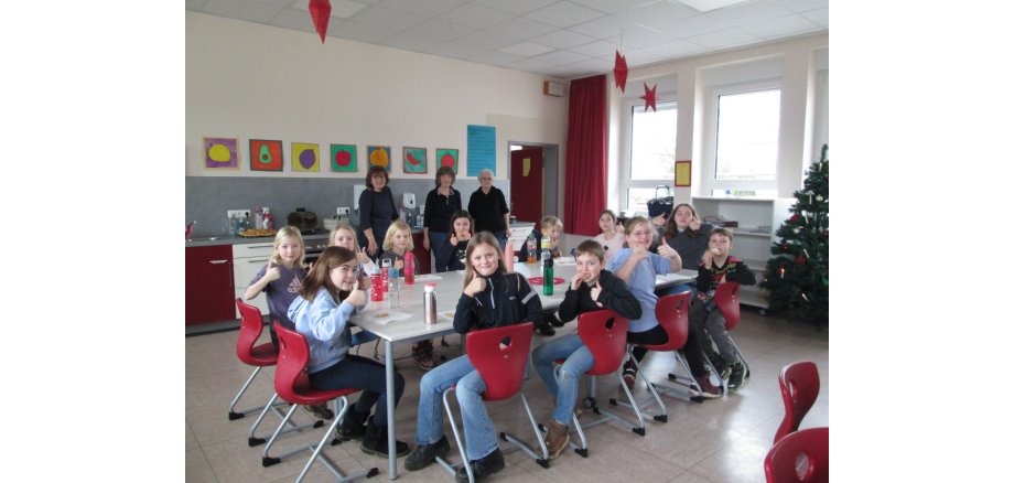 Auf dem Bild zu sehen, Kinder, die um einen Tisch sitzen; sowie die Landfrauen, die mit den Kindern Waffeln backten