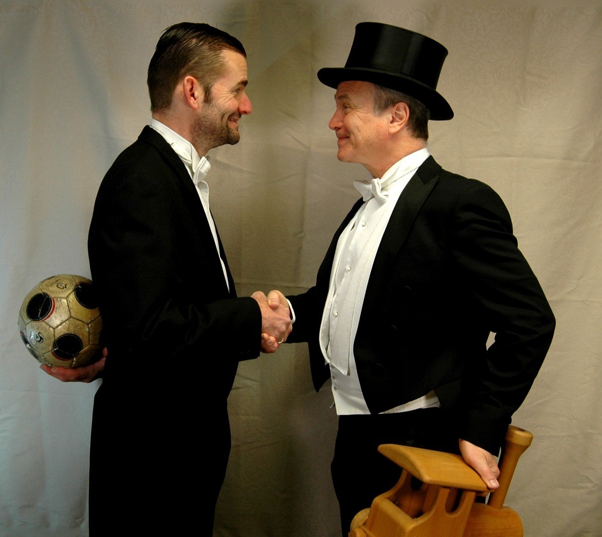 Zwei Männer mit Anzügen gekleidet, die sich gegenüber stehen und die Hand geben. der eine Mann trägt einen Zylinder und hat eine Holzeisenbahn in der anderen Hand. der andere Mann hat einen Fußbal in der Hand und hält diesen hinter seinen Rücken.