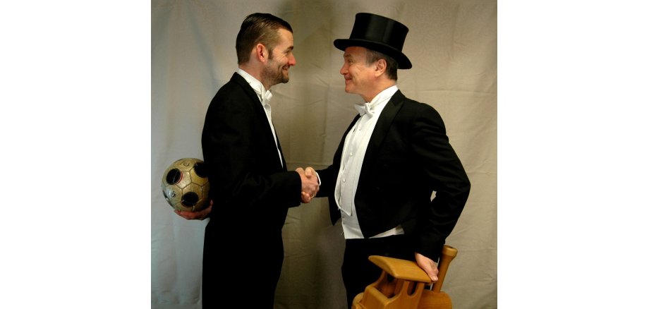 Zwei Männer mit Anzügen gekleidet, die sich gegenüber stehen und die Hand geben. der eine Mann trägt einen Zylinder und hat eine Holzeisenbahn in der anderen Hand. der andere Mann hat einen Fußbal in der Hand und hält diesen hinter seinen Rücken.