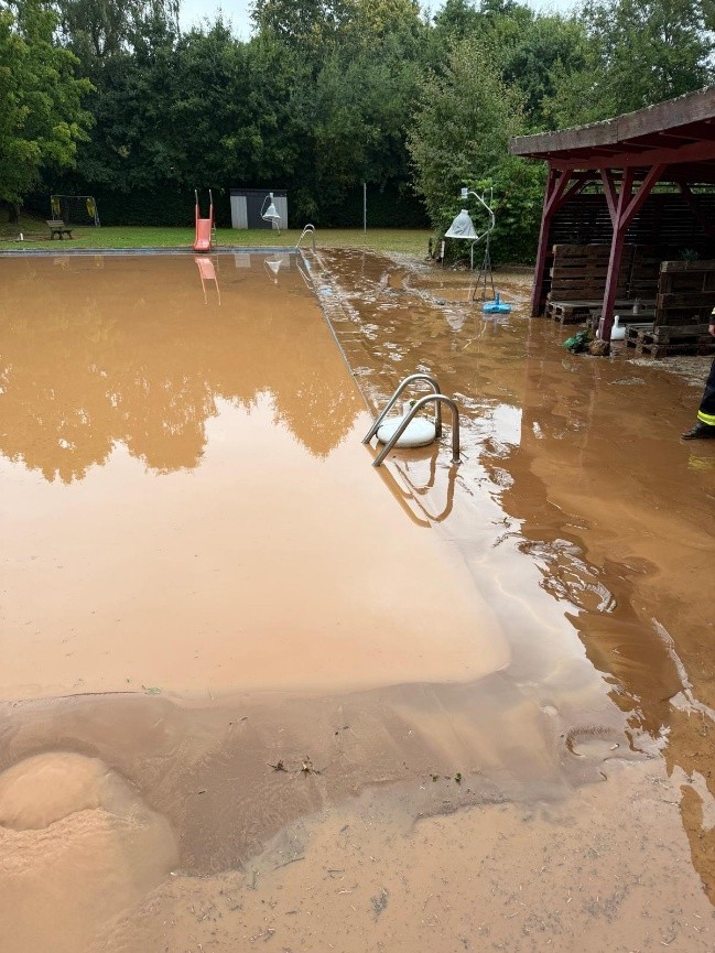 Auf dem Bild ist das Schwimmbad in Moischeid nach der Überschwemmung zu sehen; es ist übergelaufen; um das mit braunem Regenwasser gefüllte Becken ist alles mit braunem Schlamm bedeckt.