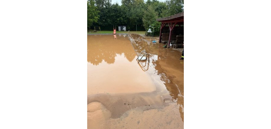 Auf dem Bild ist das Schwimmbad in Moischeid nach der Überschwemmung zu sehen; es ist übergelaufen; um das mit braunem Regenwasser gefüllte Becken ist alles mit braunem Schlamm bedeckt.