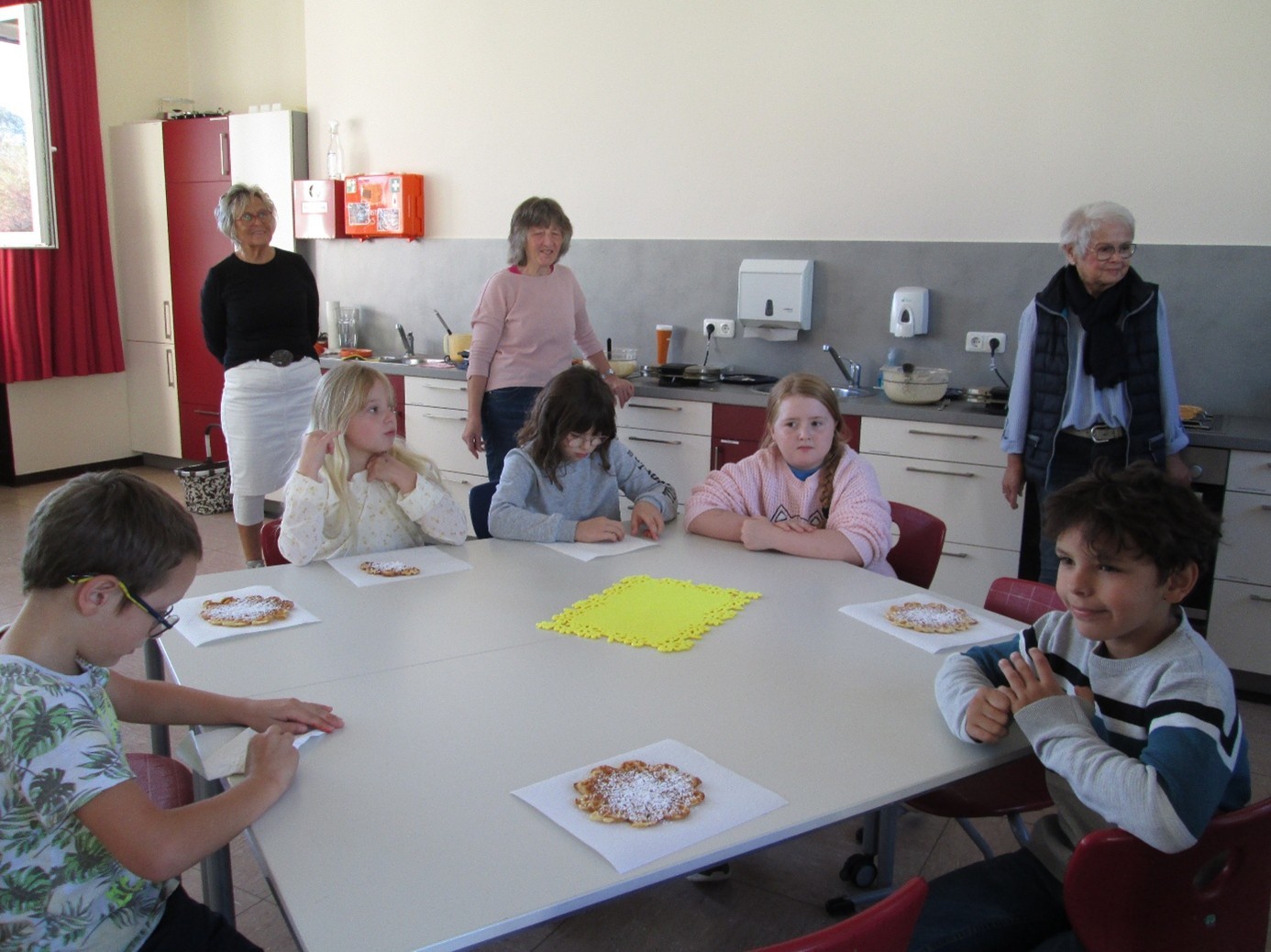 Kinder sitzen an einem Tisch und essen mit Puderzucker bestäubtes Gebäck. Drei Erwachsene stehen dahinter in einer Küche und begleiten die gemeinsame Aktivität.