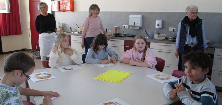 Kinder sitzen an einem Tisch und essen mit Puderzucker bestäubtes Gebäck. Drei Erwachsene stehen dahinter in einer Küche und begleiten die gemeinsame Aktivität.