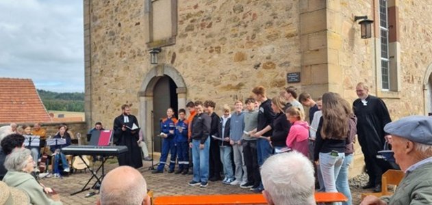 Jugendliche und Erwachsene stehen vor einer historischen Kirche und musizieren gemeinsam. Eine Gruppe von Jugendlichen steht vor dem Eingang, daneben sitzen weitere Personen an Keyboard und anderen Instrumenten