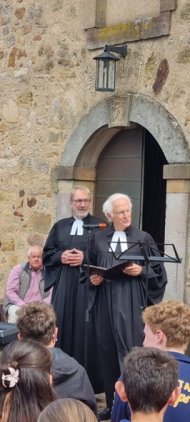 Zwei Geistliche stehen vor dem Eingang einer historischen Kirche und sprechen zu einer Gruppe von Jugendlichen. Eine Person hält ein Mikrofon, die andere ein Blatt Papier